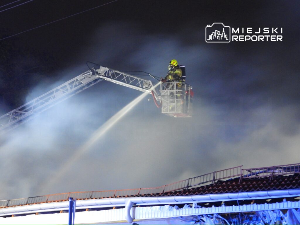 Strażak w podnośniku gaśniczym polewa wodą palący się budynek w gęstej mgle dymu nocą.