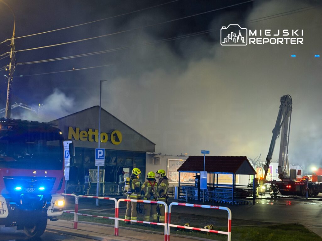 Strażacy w hełmach gaszą pożar w sklepie Netto, w tle widoczny dym i wóz strażacki z włączonymi sygnałami.