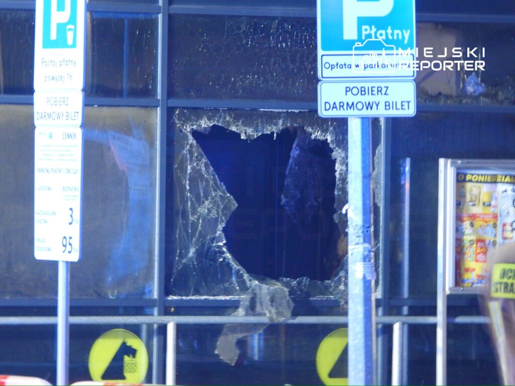 Rozbita szyba w witrynie sklepu, obok znak informacyjny o płatnym parkowaniu.