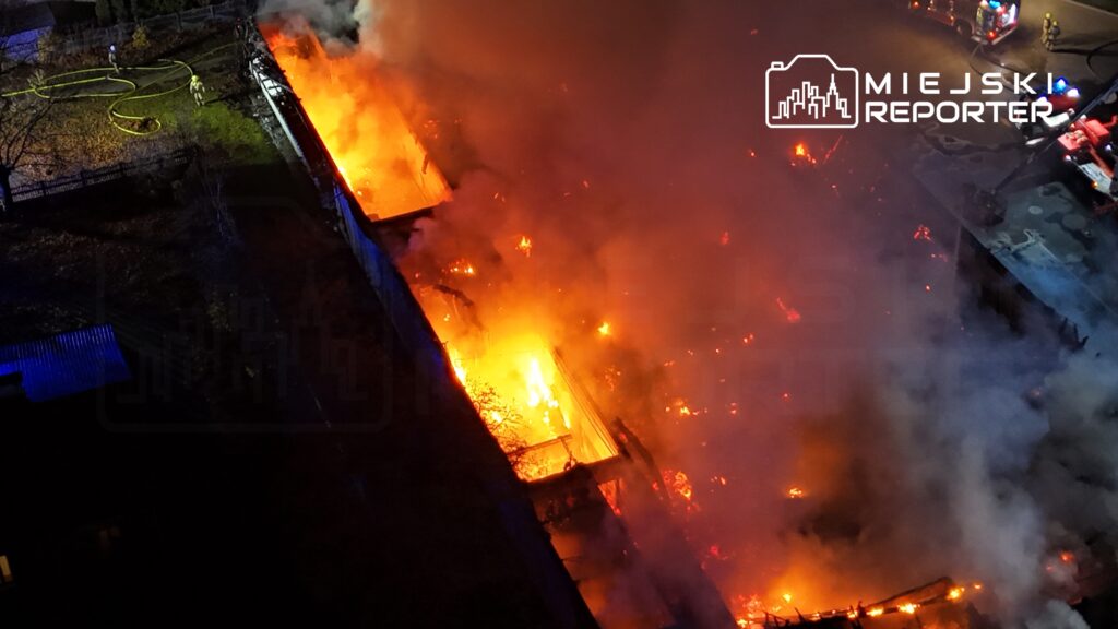 Płonący budynek otoczony gęstym dymem, w tle widać strażaków używających węży gaśniczych.