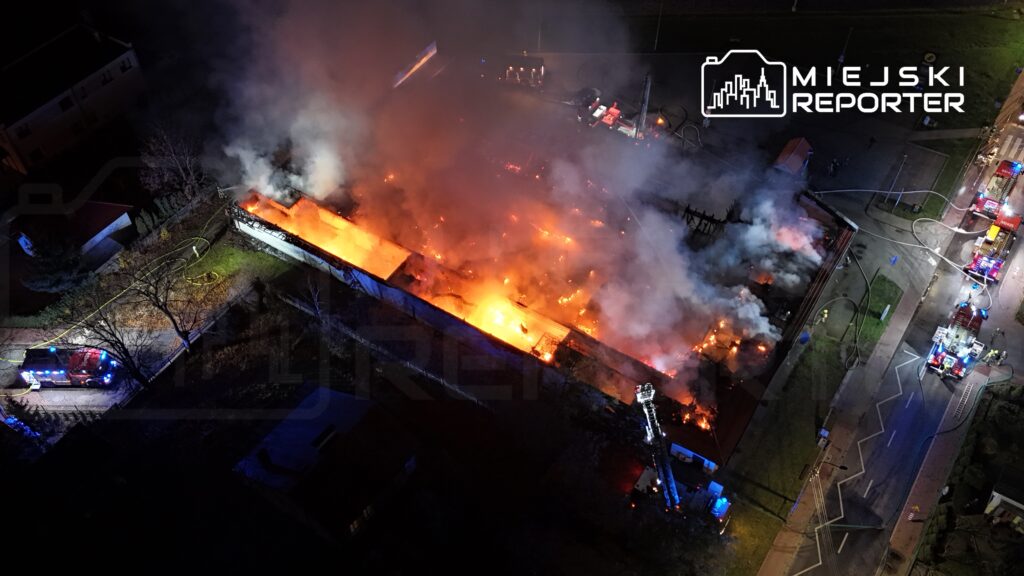 Pożar budynku przemysłowego z intensywnym dymem i płomieniami, w tle widać pojazdy straży pożarnej.
