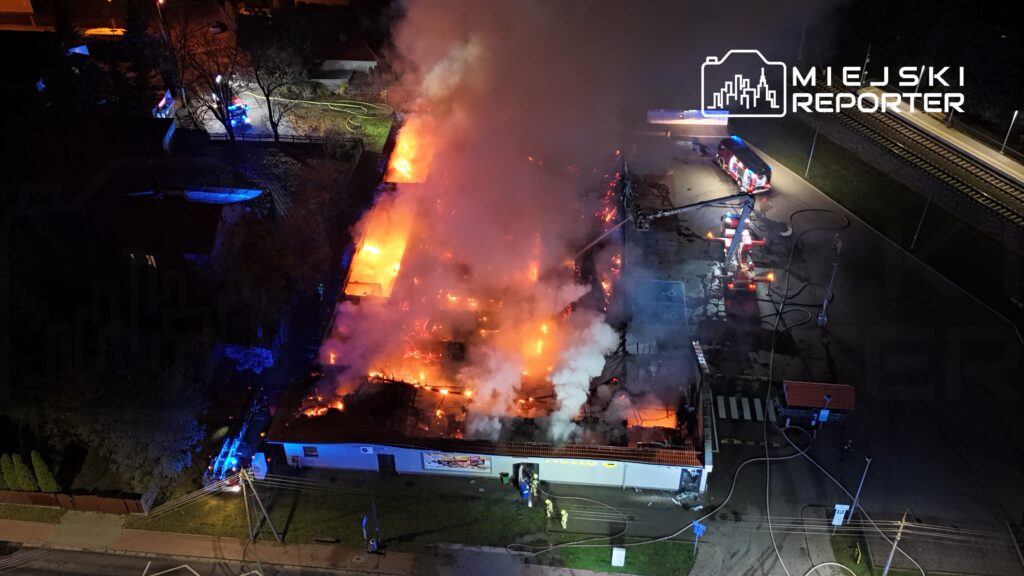 Pożar zniszczył budynek, z którego wydobywają się płomienie i gęsty dym; strażacy walczą z ogniem przy użyciu wozu strażackiego.