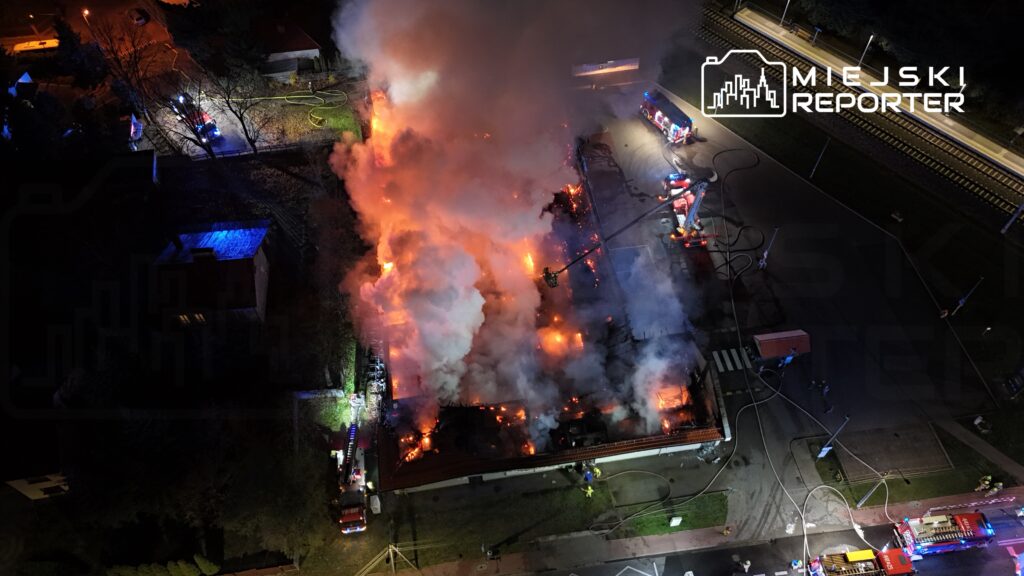 Pożar budynku z płomieniami i dymem, w tle widać wozy strażackie oraz strażaków gaszących ogień.
