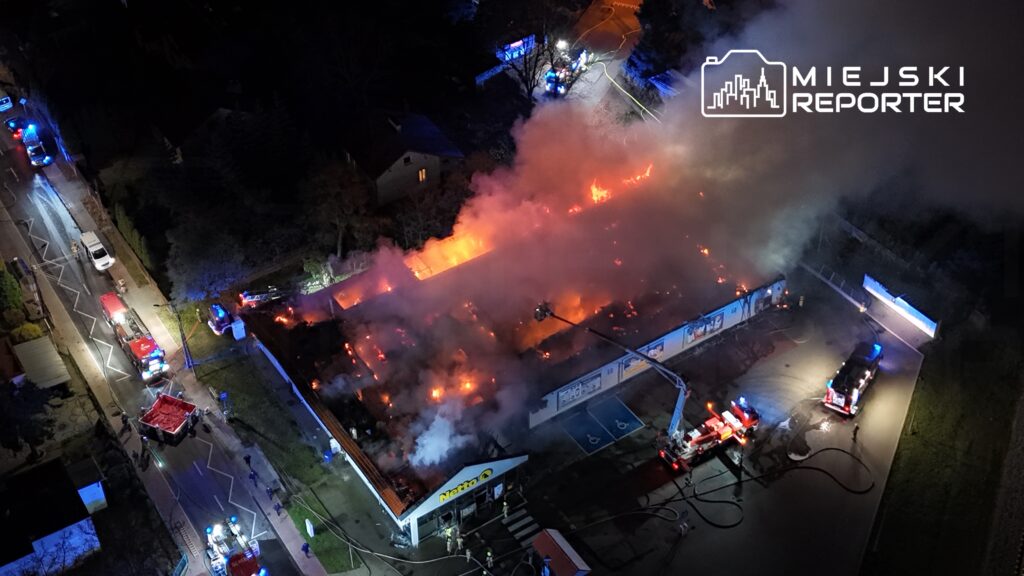 Płonący budynek handlowy, w otoczeniu wozów strażackich i dymu, w nocy. Strażacy walczą z ogniem.