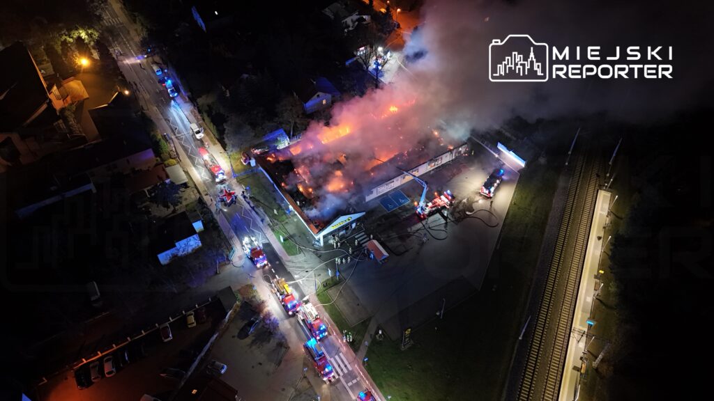 Płonący budynek z dymem unoszącym się w powietrzu, wokół liczne samochody strażackie i ratownicy.