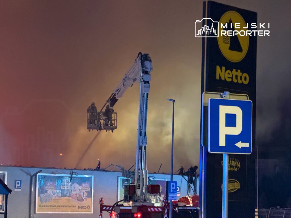 Strażacy używają podnośnika, aby gasić pożar w pobliżu budynku z logo Netto w tle.
