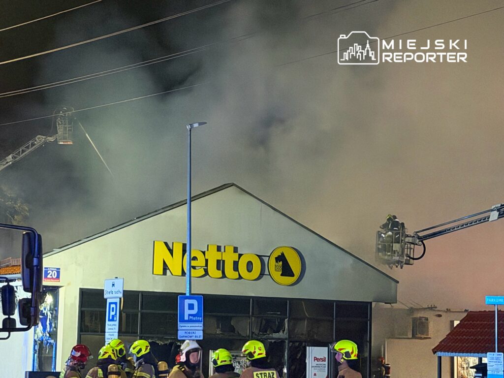 Strażacy w hełmach gaszą pożar w budynku sklepu Netto, otoczeni przez gęsty dym i sprzęt gaśniczy.