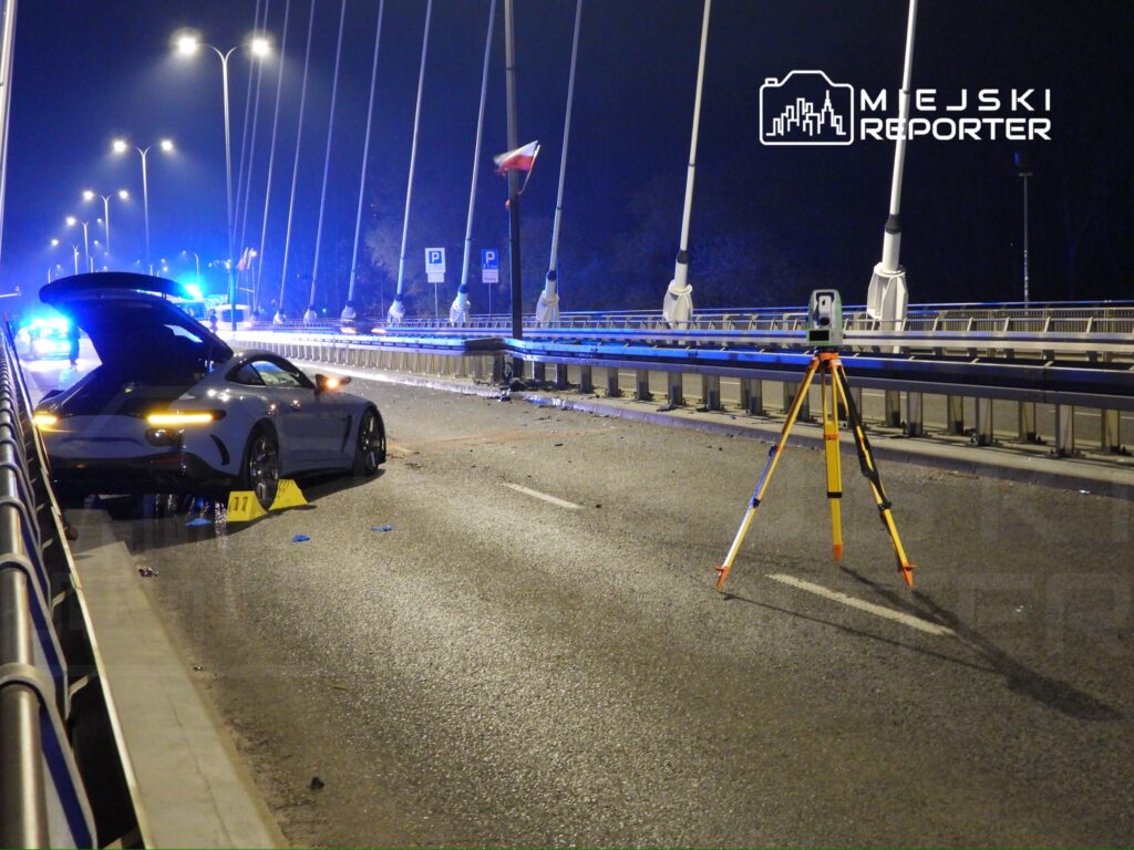 Na moście nocą stoi uszkodzony samochód sportowy z otwartą klapą, a obok znajduje się sprzęt pomiarowy.
