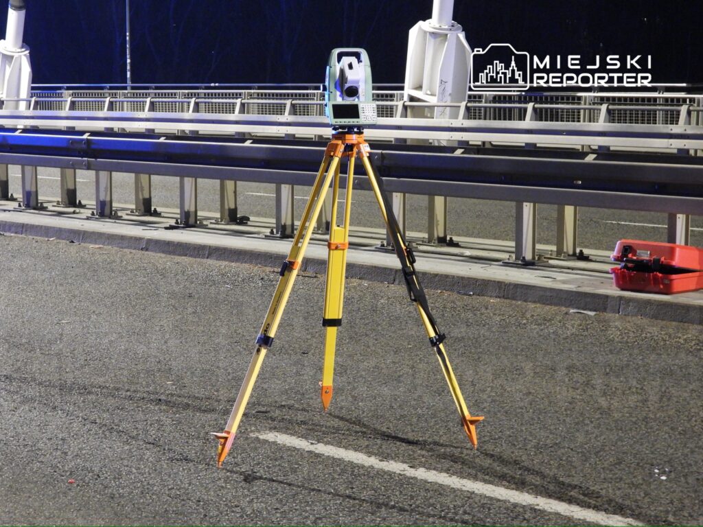 Statyw z przyrządem pomiarowym ustawiony na drodze, w pobliżu barierki ochronnej.