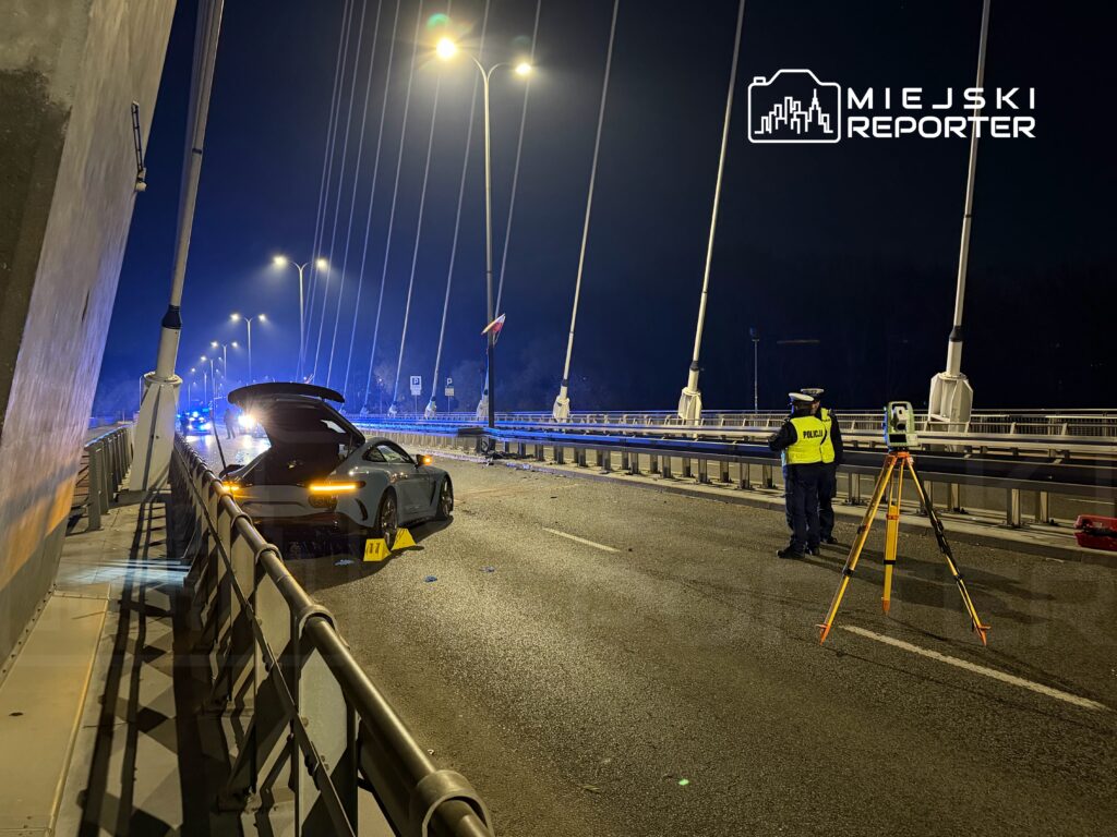 Na moście stoi policyjny funkcjonariusz, obok uszkodzony samochód sportowy z otwartym bagażnikiem.