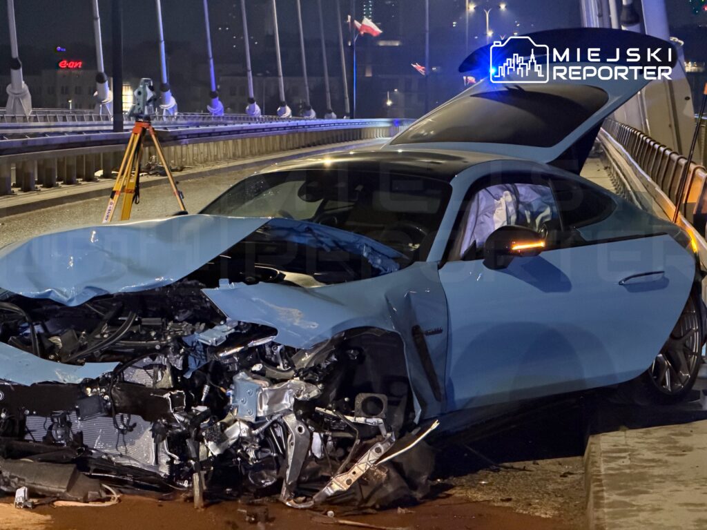 Uszkodzony niebieski samochód osobowy stoi na moście, z widocznymi uszkodzeniami przodu i otwartą klapą bagażnika.