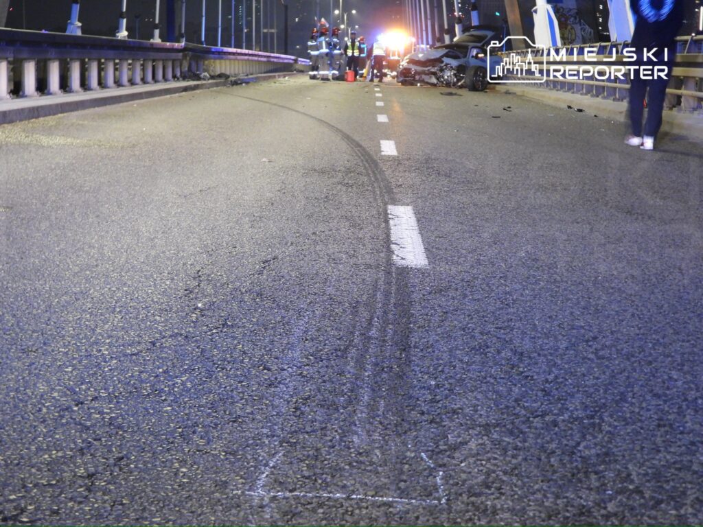 Na moście w nocy, służby ratunkowe pracują przy wypadku drogowym, obok widoczne zarysowane ślady opon na jezdni.