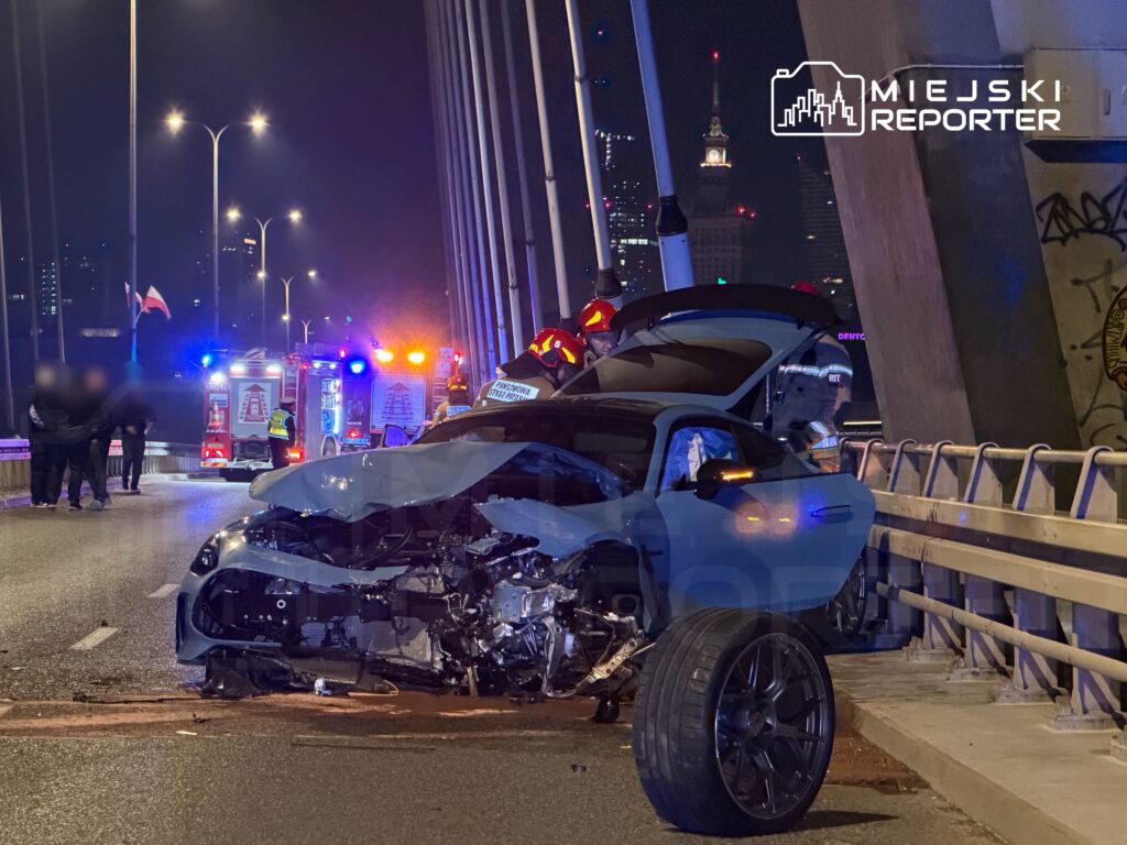 W nocy na moście stoi uszkodzony samochód osobowy, obok pracują strażacy i policja, w tle widać miejskie światła.