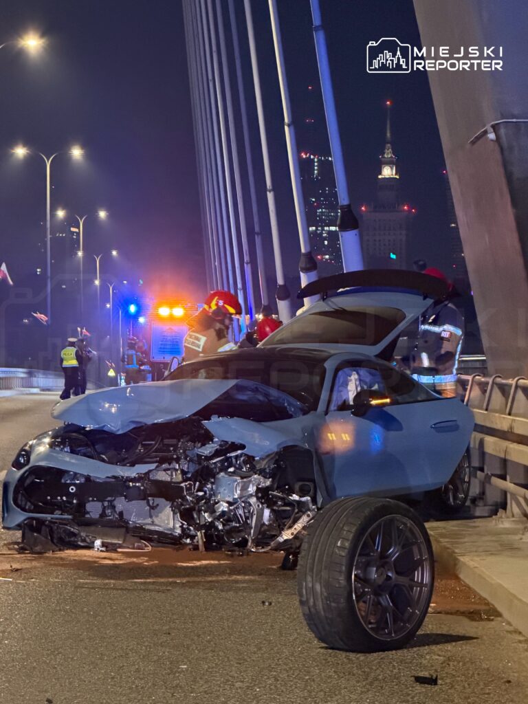 Uszkodzony samochód osobowy z odpadniętym kołem stoi na moście, obok pracują strażacy i policjanci.