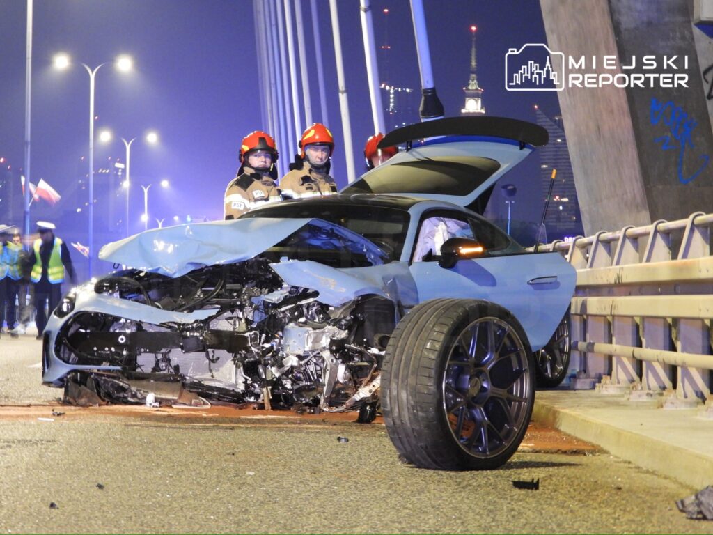 W nocy na moście stoi uszkodzony niebieski samochód sportowy, obok dwóch strażaków w hełmach.