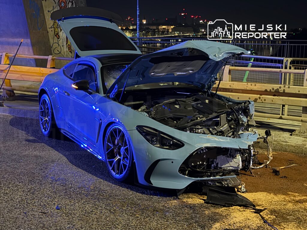 Uszkodzony samochód sportowy w jasnoniebieskim kolorze stoi na drodze, z otwartą maską i widocznymi uszkodzeniami przodu.