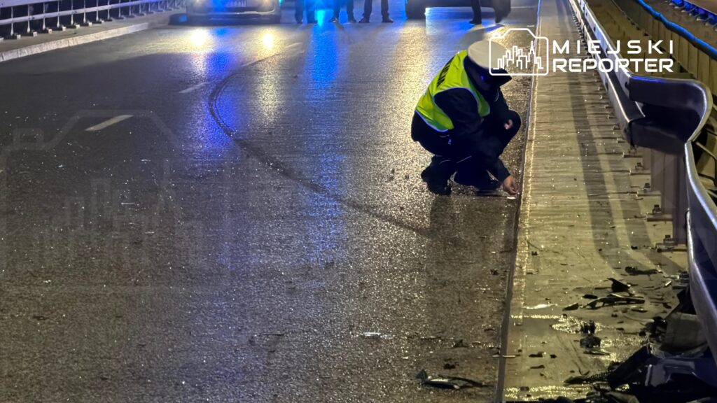 Policjant w odblaskowej kamizelce bada ślady na mokrej nawierzchni mostu po wypadku drogowym.