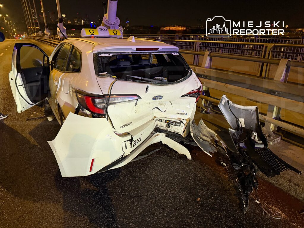 Uszkodzony samochód osobowy typu taxi z zniszczonym tyłem stoi na drodze nocą, obok otwartych drzwi.