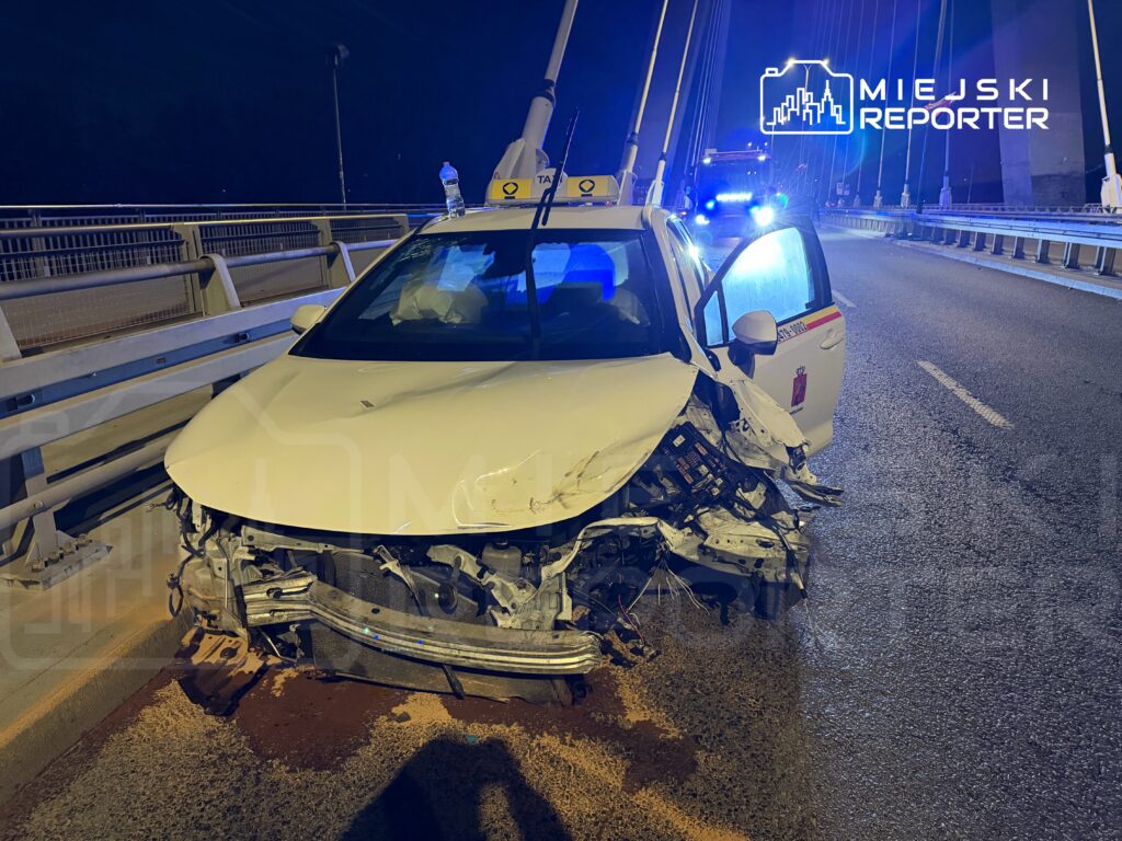 Uszkodzony samochód osobowy stoi na moście, a w tle widać niebieskie światła radiowozu.
