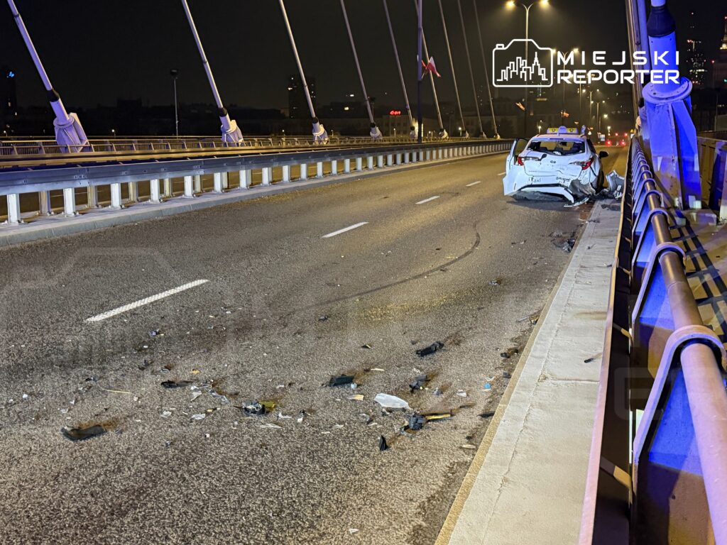 Uszkodzony taksówka leży na pasie ruchu na moście, wokół rozrzucone szczątki pojazdu i odłamki.