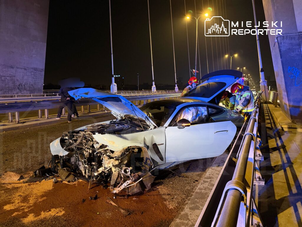 Zniszczony samochód sportowy z otwartą maską na moście, obok dwóch strażaków sprawdzających jego wnętrze.