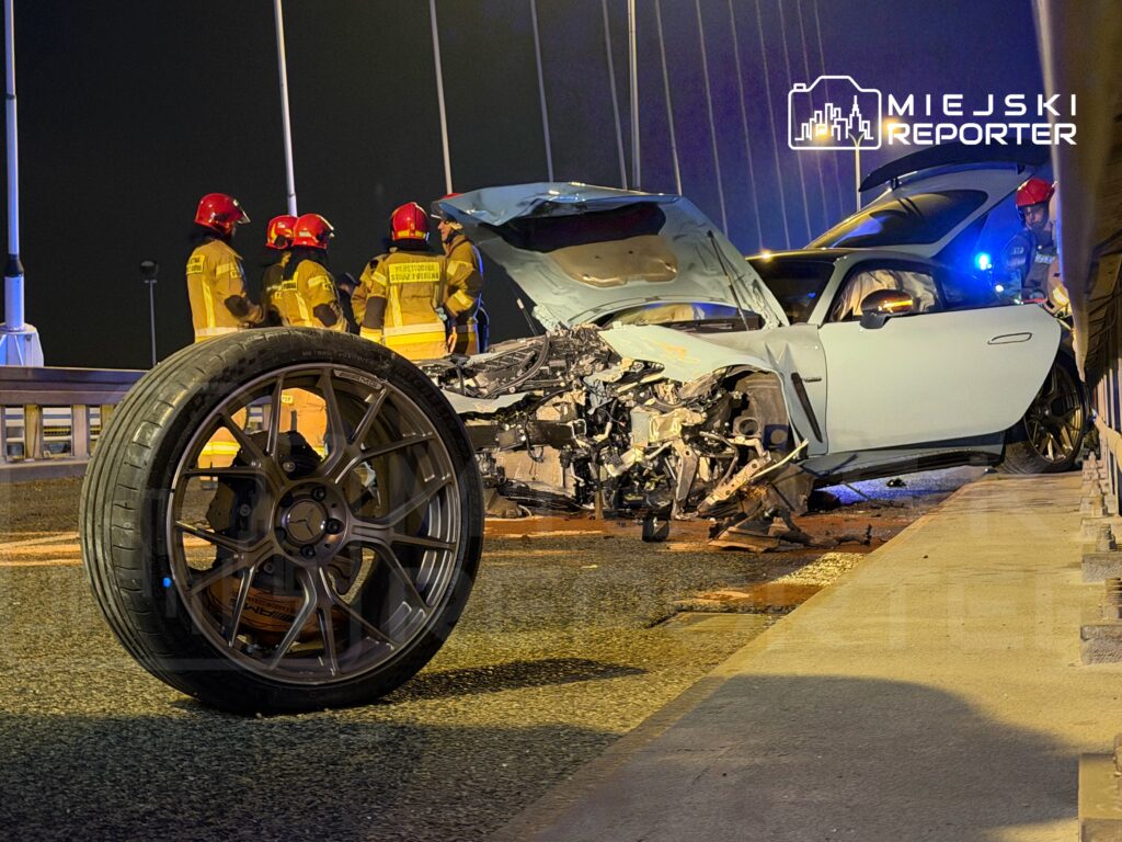W nocy na moście uszkodzony samochód sportowy z odrzuconym kołem, obok pracują strażacy w mundurach.