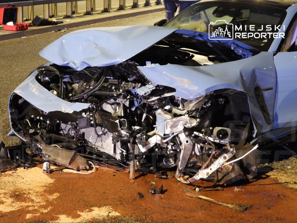 Zniszczony samochód osobowy leży na jezdni, z widocznymi uszkodzeniami przodu i rozlanym olejem.