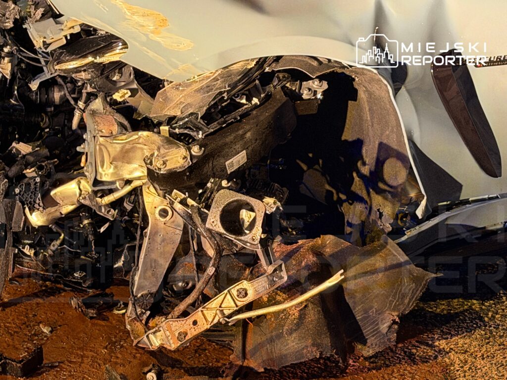 Zdeformowany przód samochodu po wypadku, z widocznymi uszkodzeniami silnika i zniszczonym podwoziem, na mokrej nawierzchni.