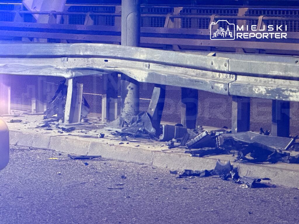 Zniszczona bariera ochronna oraz fragmenty pojazdu na poboczu drogi, w tle niebieskie światła policyjne.