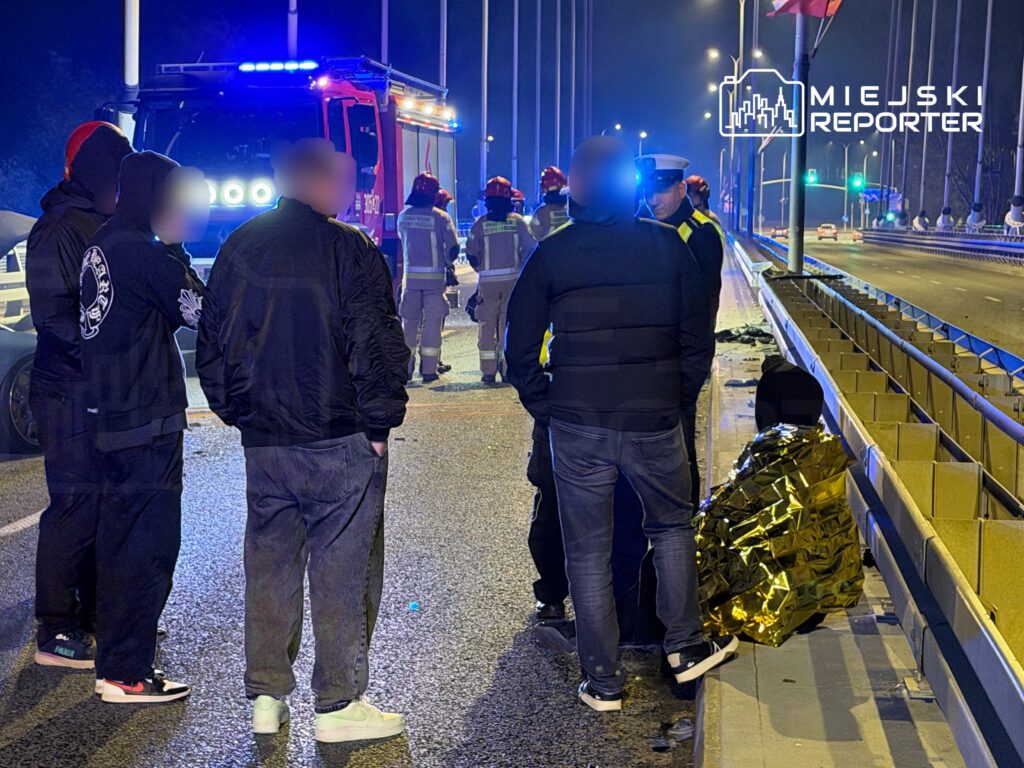 Grupa osób w ciemnych ubraniach obserwuje działania strażaków na oświetlonym moście, gdzie znajduje się wóz strażacki. Na nawierzchni leżą fragmenty pojazdu.