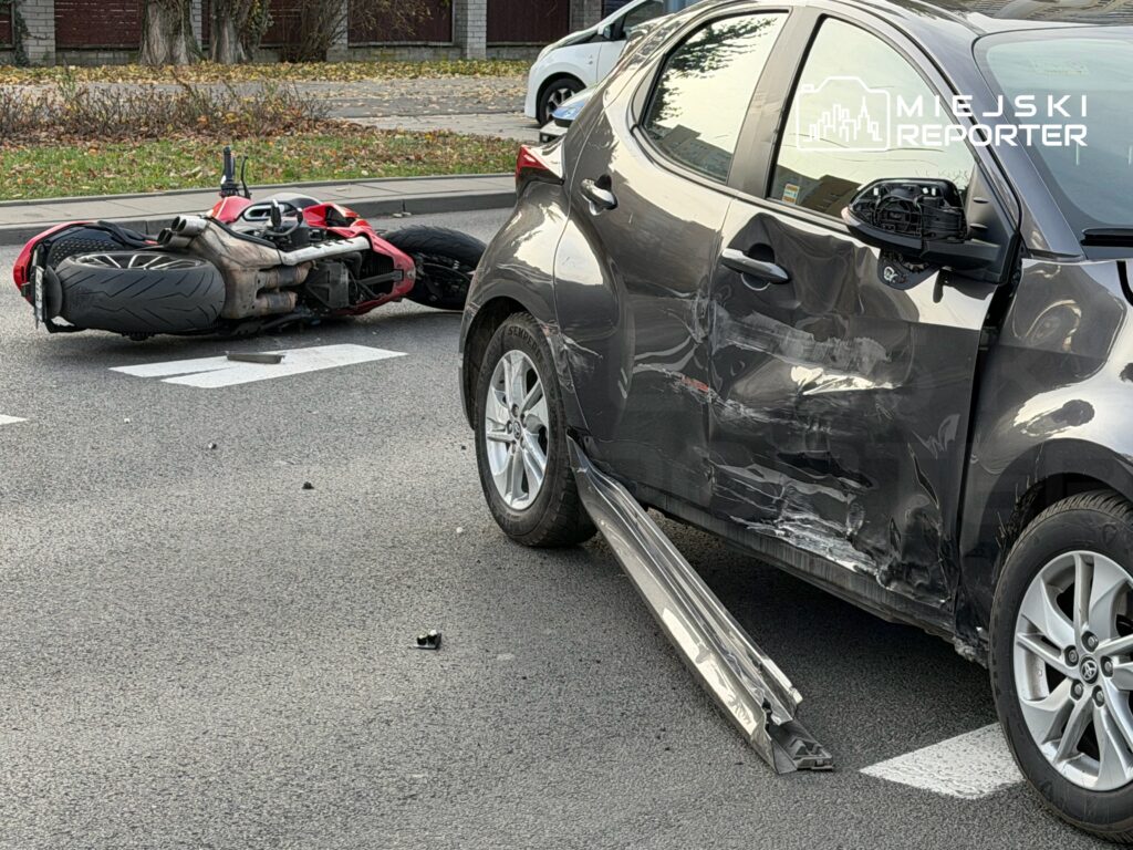 Motocykl leży na boku na jezdni obok uszkodzonego samochodu osobowego z wgniecioną boczną częścią.