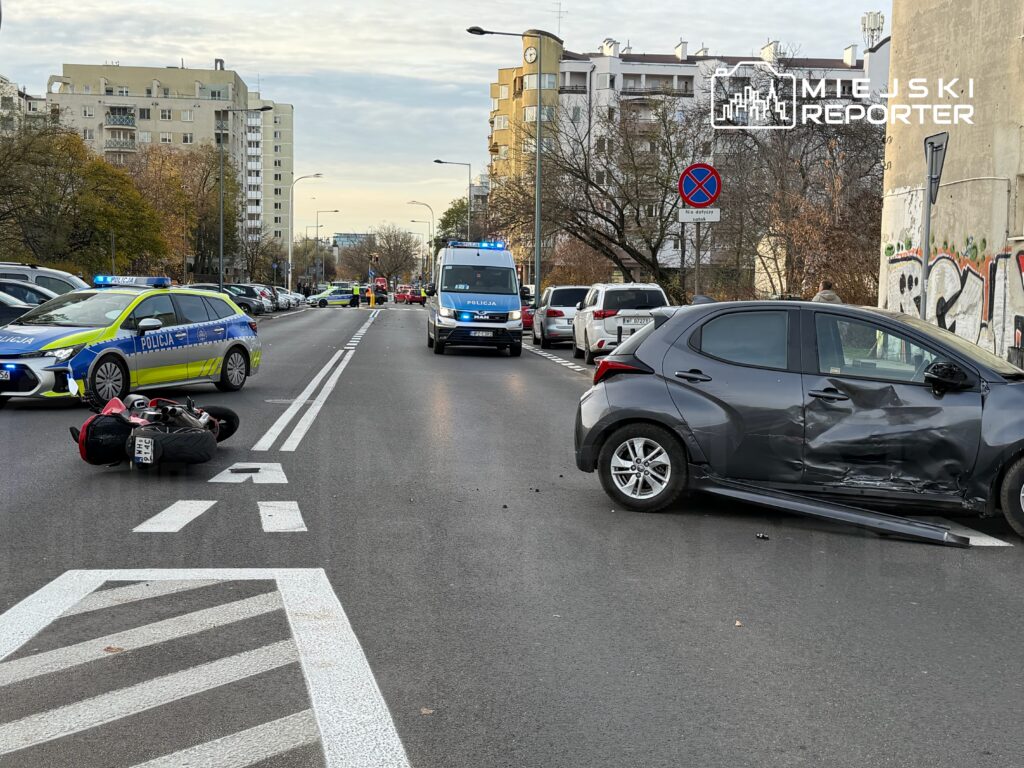 Motocykl leży na drodze obok uszkodzonego samochodu, a w tle widoczne są radiowozy policyjne.