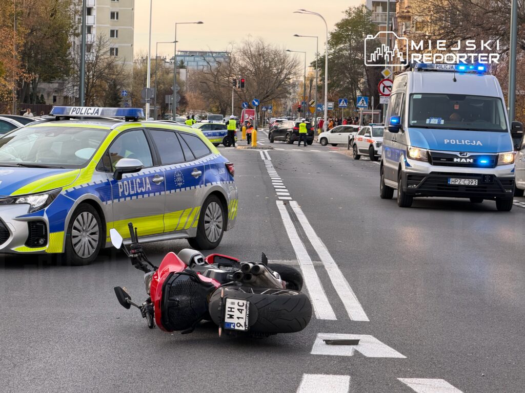 Motocykl leży na jezdni obok radiowozu policyjnego, a na drodze widać funkcjonariuszy zabezpieczających miejsce zdarzenia.