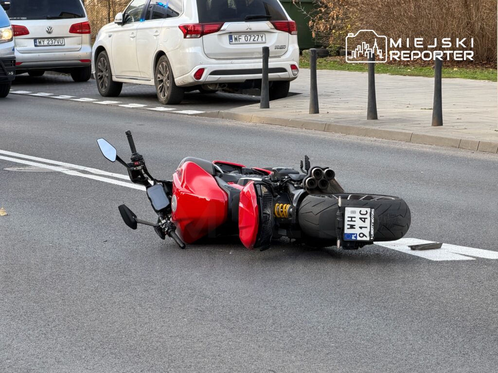 Czerwony motocykl leży na boku na jezdni, obok zaparkowanych samochodów, w miejskim otoczeniu.