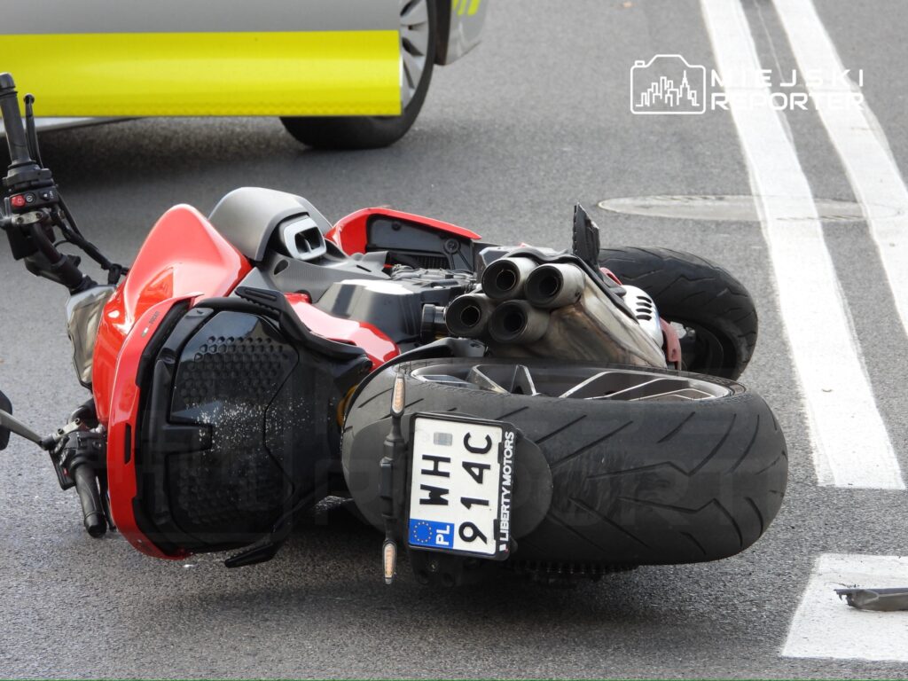 Czerwony motocykl leży na boku na jezdni, obok widać samochód z widoczną częścią nadwozia.