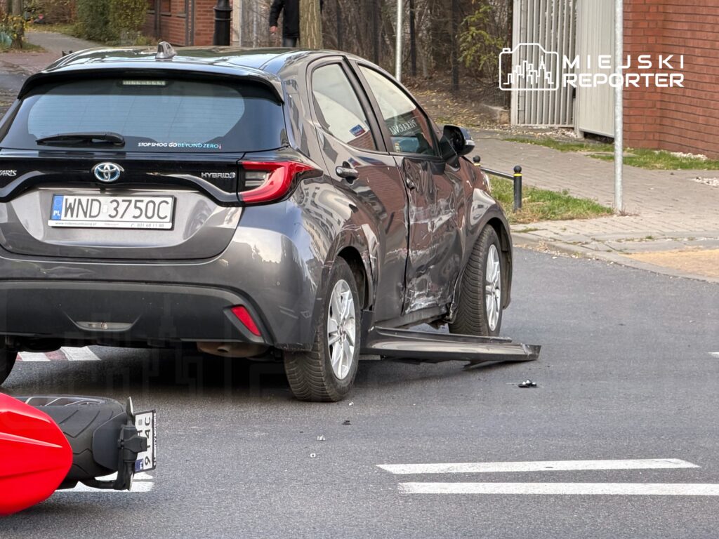 Uszkodzony samochód osobowy stojący na skrzyżowaniu, obok leży fragment motocykla.
