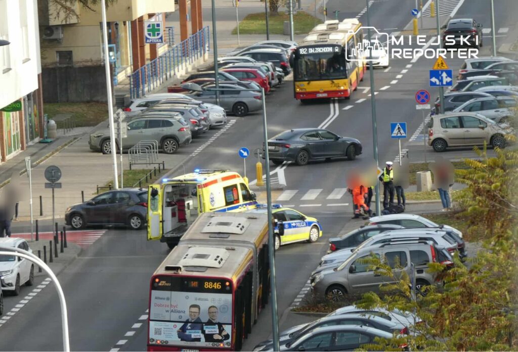 Na skrzyżowaniu zatrzymane pojazdy, w tym karetka i radiowóz, obok grupka osób w odblaskowych kamizelkach.