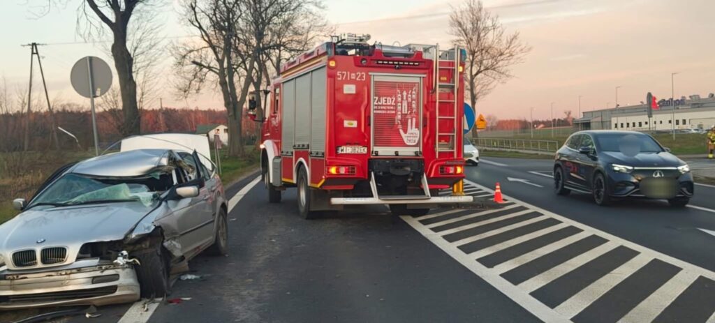 Zniszczony samochód osobowy zaparkowany na poboczu drogi, obok stoi wóz strażacki z włączonymi światłami.