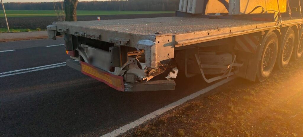 Uszkodzona tylna część naczepy ciężarówki zaparkowanej na poboczu drogi, w tle widoczne pola i drzewa.