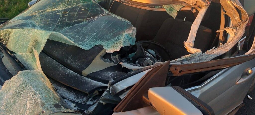 Zniszczony samochód z rozbitą szybą czołową i uszkodzonym wnętrzem, leżący na poboczu drogi.