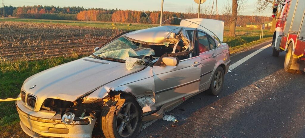 Uszkodzony samochód osobowy leży na poboczu drogi, obok niego stoi wóz strażacki, a w tle widać pola i drzewa.