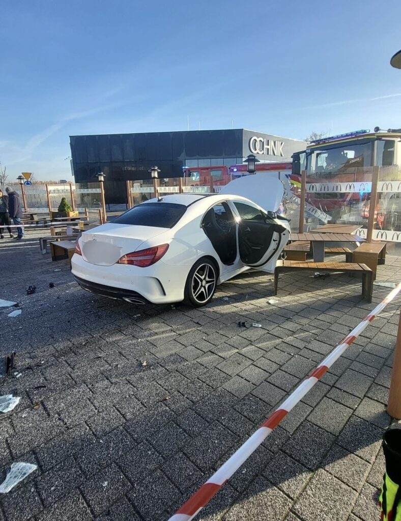 Biały samochód osobowy stoi z otwartymi drzwiami na parkingu, w tle widoczne budynki i woz strażacki.