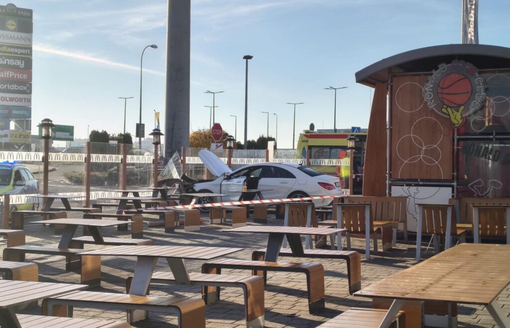 Biały samochód osobowy z otwartą maską stoi na parkingu obok stolików w strefie gastronomicznej, w tle widać karetkę.