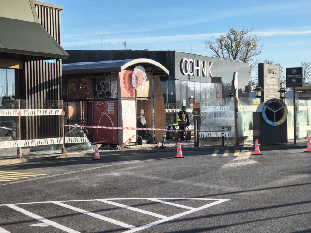 Robotnicy w kamizelkach odblaskowych naprawiają uszkodzoną konstrukcję w pobliżu restauracji fast food z przeszkloną fasadą.