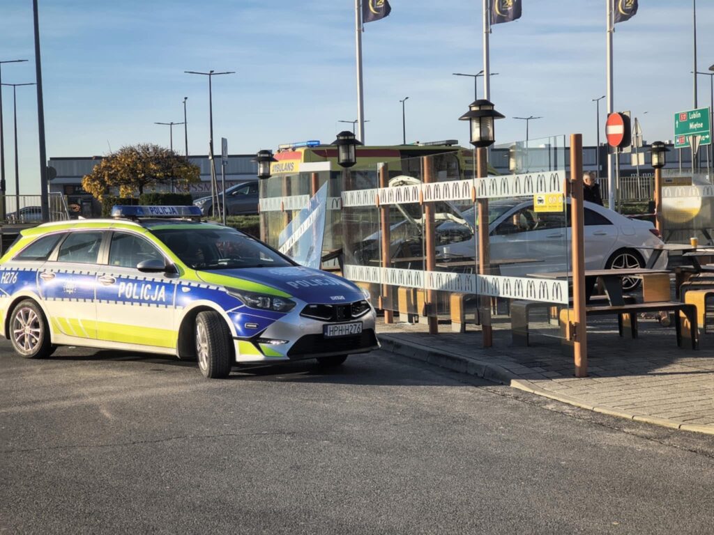 Policyjny radiowóz zaparkowany obok restauracji fast food, w tle widoczne inne pojazdy i znaki drogowe.