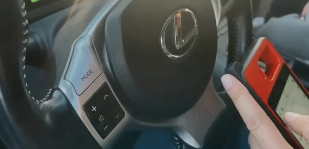 Kierownica samochodu z logo Lexus, ręka trzymająca telefon w czerwonym etui, przyciski sterujące w zasięgu ręki.