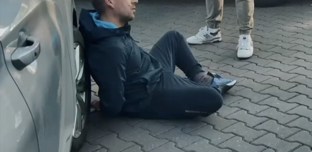 Mężczyzna w ciemnej odzieży siedzący na chodniku obok samochodu, z nogami wyciągniętymi w stronę przechodnia.