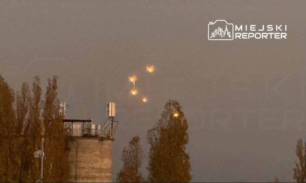 Oświetlone obiekty poruszające się po niebie nad silosami, w tle drzewa i szare niebo.