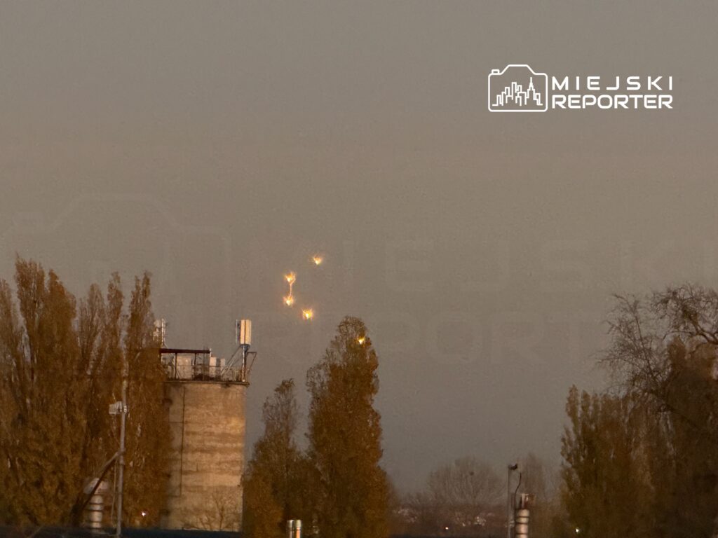 Świecące obiekty na niebie nad przemysłowym terenem z silosami i drzewami w tle.