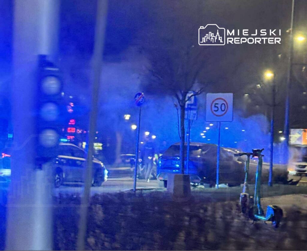 Nocna scena na ulicy z niebieskimi światłami policyjnymi, zaparkowanym samochodem i elektryczną hulajnogą.